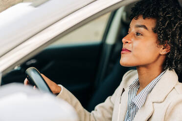 Seitenansicht einer fröhlichen afroamerikanischen Fahrerin in modischem Outfit, die lächelnd ein modernes Auto auf der Straße fährt - ADSF24784