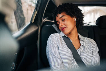 Sorglose junge Afroamerikanerin mit TWS-Kopfhörern, die mit geschlossenen Augen Musik hört und sich in einem modernen Auto ausruht - ADSF24786