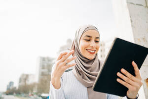 Inhalt Muslimische Frau mit Kopftuch, die mit einem Getränk zum Mitnehmen in der Nähe eines Brunnens in der Stadt sitzt und auf einem Tablet surft, während sie in die Kamera schaut - ADSF24790