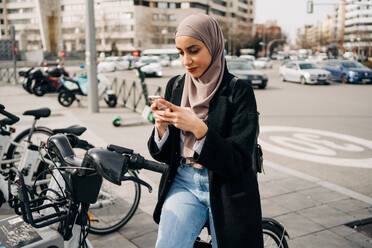 Side view of Muslim female in headscarf using bicycle sharing system in city - ADSF24793