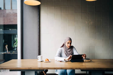 Content Muslim female in hijab and talking on video chat via tablet while sitting at table in cafe - ADSF24802