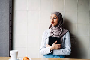 Seitenansicht einer muslimischen Freiberuflerin mit traditionellem Hidschab, die in einem Café steht und mit einem Tablet an einem Projekt arbeitet und dabei wegschaut - ADSF24807