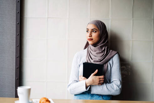 Side view of Muslim female freelancer wearing traditional hijab standing in cafe with tablet while working on project remotely and looking away - ADSF24807
