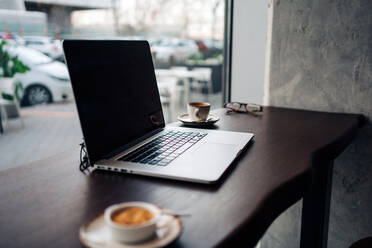 Modernes Netbook mit schwarzem Bildschirm auf einem Holztisch mit Tassen mit aromatischem Kaffee in einer Cafeteria - ADSF24814