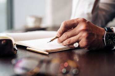 Crop anonymous businessman in wristwatch with pen and open journal working at cafeteria table in daylight - ADSF24819