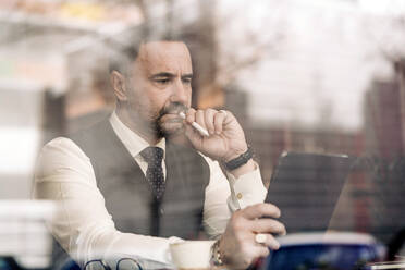 Thoughtful middle aged ethnic male entrepreneur touching mouth with stylus while using tablet behind glass wall in cafeteria - ADSF24826