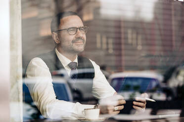 Thoughtful middle aged ethnic male entrepreneur using tablet behind glass wall in cafeteria - ADSF24827