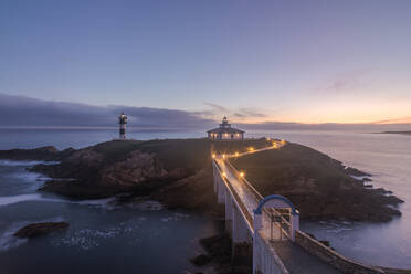 Spektakuläre Landschaft der Brücke, die zu felsigen Insel mit grünem Gras mit Leuchtturm in welligen Ozean in Faro Illa Pancha in Galicien in Spanien in der Dämmerung platziert bedeckt - ADSF24852