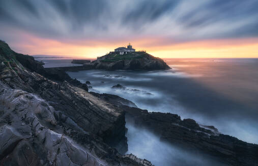 Breathtaking scenery of rocky island with lighthouse located in ocean near rocky coast in Faro Tapia de Casariego in Asturias in Spain under cloudy sky at sunrise - ADSF24857