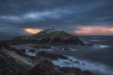 Atemberaubende Landschaft der felsigen Insel mit Leuchtturm im Meer in der Nähe von felsigen Küste in Faro Tapia de Casariego in Asturien in Spanien unter bewölktem Himmel bei Sonnenaufgang befindet - ADSF24858