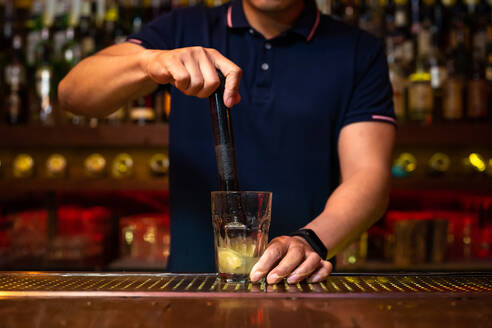 Unrecognizable bartender crushing lemon wedges in the glass while preparing mojito cocktail in the bar - ADSF24872