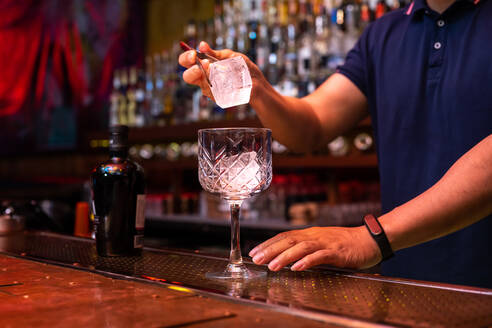 Unrecognizable bartender putting a big ice cube into the glass while preparing a gin tonic cocktail in the bar - ADSF24876