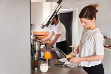 Seitenansicht einer ruhigen jungen Frau in einem weißen T-Shirt, die ein gesundes Frühstück mit Avocado zubereitet, während sie am Tisch in der Küche steht, in der Nähe eines nicht erkennbaren Freundes, der das Geschirr spült - ADSF24889