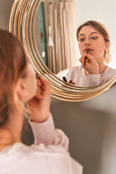 Crop young female with blond hair in casual clothes looking in mirror and applying lip liners in sunny morning - ADSF24896