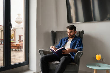 Konzentrierter junger spanischer Mann in Freizeitkleidung sitzt in einem bequemen Sessel und liest ein interessantes Buch in der Nähe des Tisches mit einem Glas frischem Orangensaft im Tageslicht zu Hause - ADSF24899