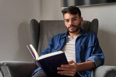 Concentrated young Hispanic guy in casual clothes sitting in comfortable armchair and reading interesting book and looking at camera in daylight at home - ADSF24900