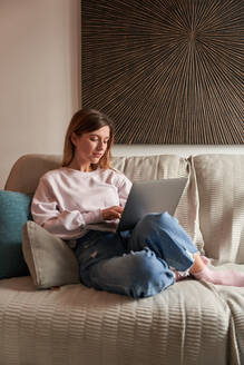 Full body of peaceful young female freelancer in casual clothes sitting on cozy sofa and working remotely on laptop - ADSF24901