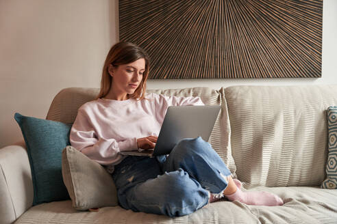 Full body of peaceful young female freelancer in casual clothes sitting on cozy sofa and working remotely on laptop - ADSF24902