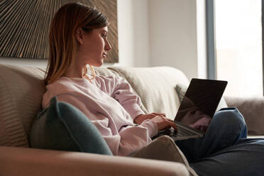 Peaceful young female freelancer in casual clothes sitting on cozy sofa and working remotely on laptop - ADSF24903