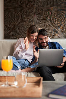 Fröhliches junges gemischtrassiges Paar in Freizeitkleidung, das lächelt und mit den Händen winkt, während es auf dem Sofa sitzt und ein Videogespräch über einen Laptop führt - ADSF24904