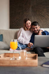 Fröhliches junges gemischtrassiges Paar in legerer Kleidung, das lächelnd auf dem Sofa sitzt und ein Videogespräch über einen Laptop führt - ADSF24905