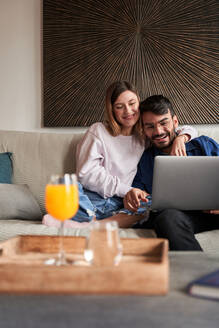 Fröhliches junges gemischtrassiges Paar in legerer Kleidung, das lächelnd auf dem Sofa sitzt und ein Videogespräch über einen Laptop führt - ADSF24905