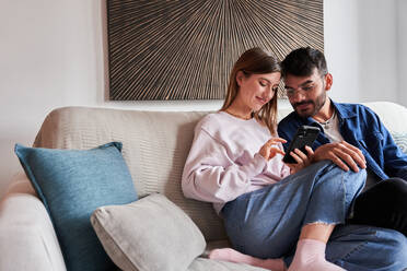 Romantisches junges Paar in legerer Kleidung, das sich auf dem Sofa entspannt und Fotos auf dem Smartphone anschaut, während des Wochenendes zu Hause - ADSF24907