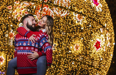 Mann nimmt Frau vor beleuchteter Weihnachtsdekoration huckepack - LJF02186