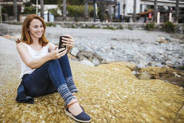 Lächelnde Frau, die ein Selfie mit ihrem Smartphone macht, während sie auf dem Gehweg sitzt - LJF02188