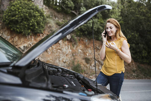 Frau, die mit einem Mobiltelefon telefoniert, um Pannenhilfe zu erhalten, während sie am Auto steht - LJF02190