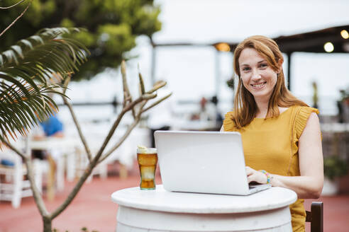 Smiling woman with laptop and drink sitting at bar - LJF02202