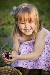 Smiling girl looking away holding cherries while crouching in backyard - ZEDF04241