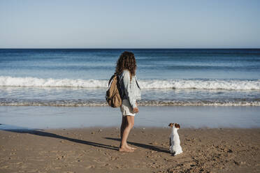 Weiblicher Rucksacktourist mit Hund am Strand bei Sonnenuntergang - EBBF04047