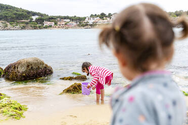 Mädchen betrachtet Schwester, die mit Eimer am Strand spielt - GEMF04869