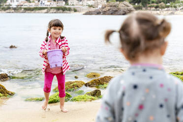 Lächelndes Mädchen mit Eimer, das seine Schwester in den Ferien am Strand betrachtet - GEMF04870