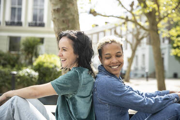 Female friends smiling while sitting back to back - PMF01845