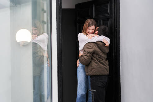 Fröhliche junge Frau trifft anonymen Freund mit Gepäck, der von einer Geschäftsreise zurückkehrt, vor der Haustür und umarmt ihn - ADSF24912