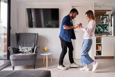 Side view of excited young multiracial couple in stylish clothes holding hands while dancing together in modern apartment in daytime - ADSF24913