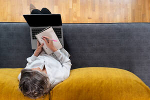 Von oben anonyme weibliche Fernarbeiterin mit Notizblock und Stift auf der Couch sitzend vor einem Netbook mit Diagrammen auf dem Bildschirm - ADSF24934