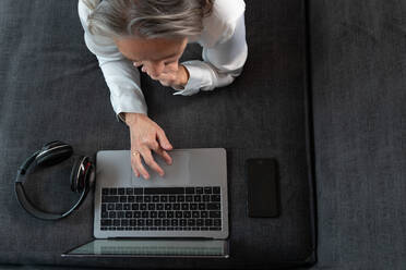 Draufsicht auf eine anonyme Fernarbeiterin, die mit einem Netbook auf der Couch mit einem drahtlosen Headset zu Hause im Internet surft - ADSF24935