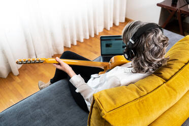 High angle back view of unrecognizable female guitarist in headphones playing musical instrument on couch with netbook at home - ADSF24939