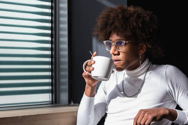 Relaxed young black guy with Afro hairstyle in stylish clothes drinking coffee and looking away while sitting on cafe terrace in city - ADSF24942