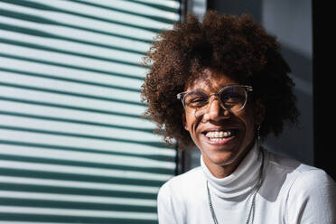 Cheerful young charismatic black male millennial with Afro hair in casual outfit and eyeglasses smiling and looking at camera sitting near window with jalousie - ADSF24944