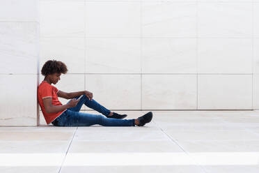 Side view of serious young African American guy with curly hair in casual clothes browsing mobile phone while sitting on tiled floor in modern building - ADSF24947