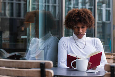 Ruhiger junger Afroamerikaner mit lockigem Haar in trendigem Outfit, der sich mit einer Tasse Kaffee auf der Terrasse eines Cafés ausruht und ein interessantes Buch liest - ADSF24948