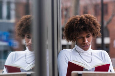 Ruhiger junger Afroamerikaner mit lockigem Haar in trendigem Outfit, der sich auf der Terrasse eines Cafés ausruht und ein interessantes Buch liest - ADSF24949