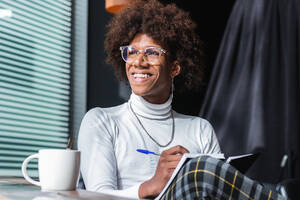 Fröhlicher junger afroamerikanischer Student mit lockigem Haar in stilvollem Outfit und Brille, der in einem modernen Café an einem sonnigen Tag Notizen in ein Heft schreibt, während er sich auf eine Prüfung vorbereitet - ADSF24953