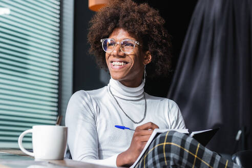 Cheerful young African American male student with curly hair in stylish outfit and eyeglasses taking notes in copybook while preparing for exam in modern cafe on sunny day - ADSF24953