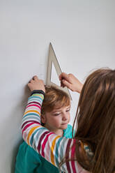 Schwester hilft Bruder beim Messen seiner Größe mit Lineal und Bleistift an der Wand - ADSF24968