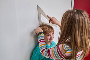 Schwester hilft Bruder beim Messen seiner Größe mit Lineal und Bleistift an der Wand - ADSF24969